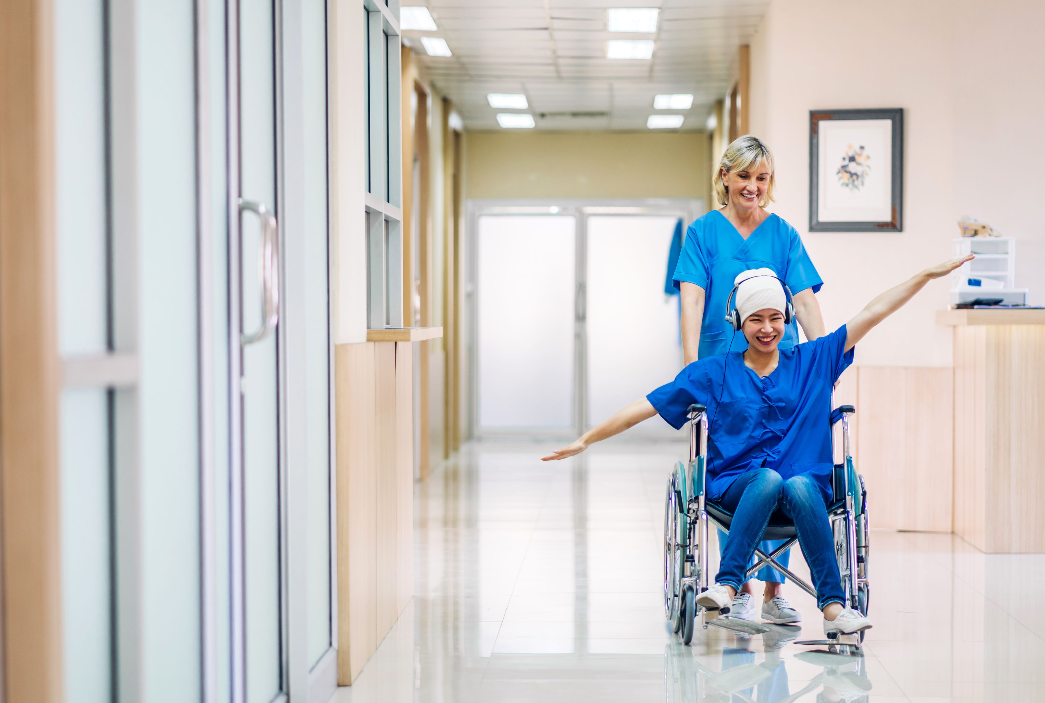 A happy female cancer patient in a wheelchair with her arms outstretched like wings being pushed through a hospital by a smiling female nurse