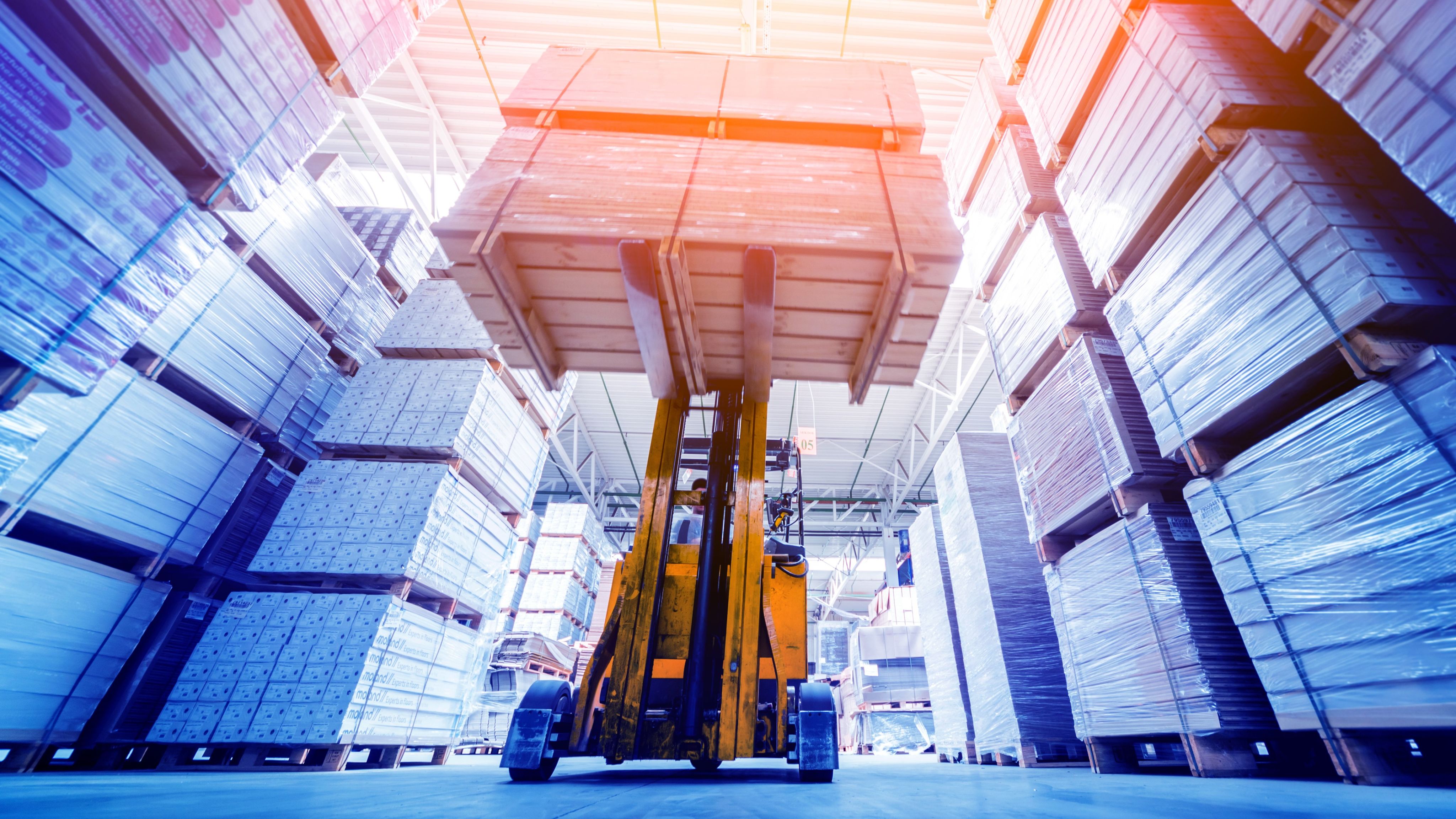A photograph showing a yellow forklift truck operating in a warehouse full of stacked pallets