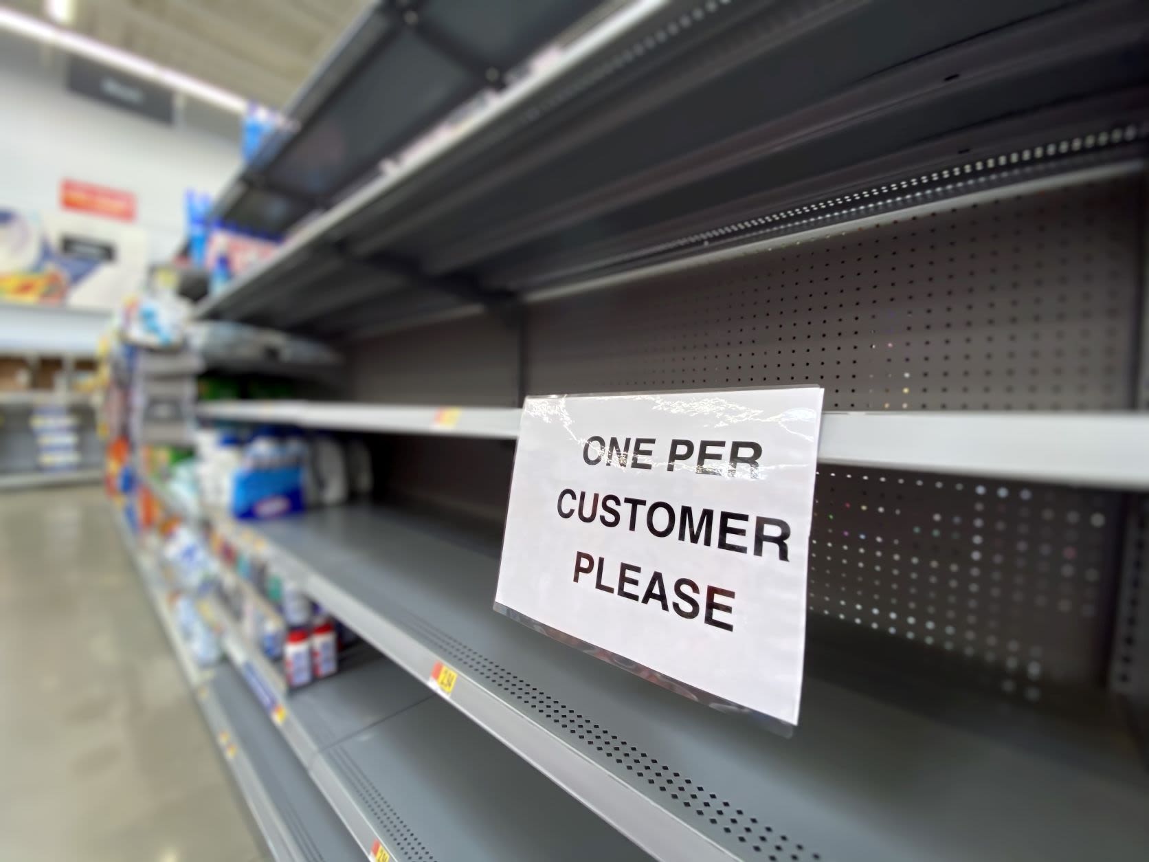 A photograph of empty supermarket shelves with a sign saying 'One per customer please'