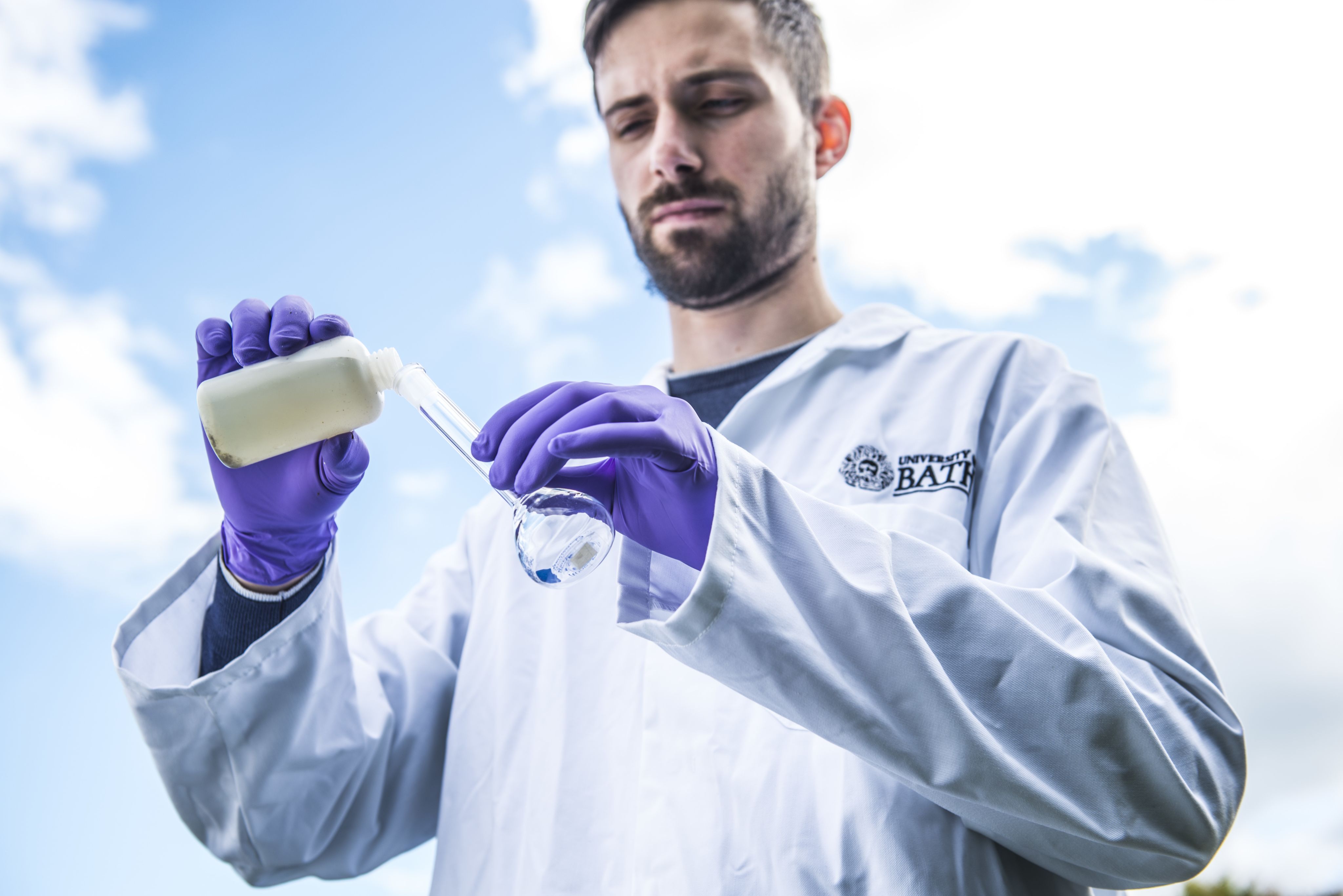Scientist in a labcoat against a cloudy sky pouring water from a bottle into a flask