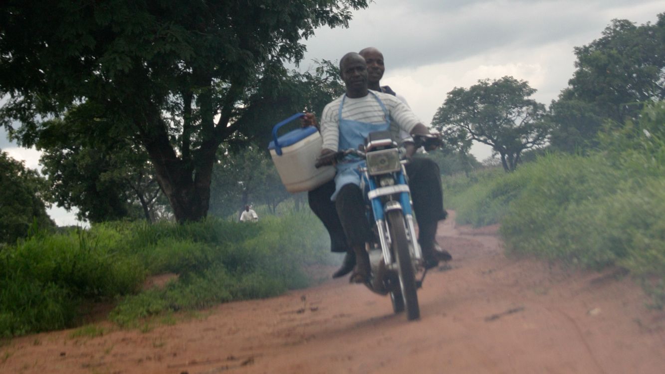 Men on motorbike drive through rural setting with vaccine box