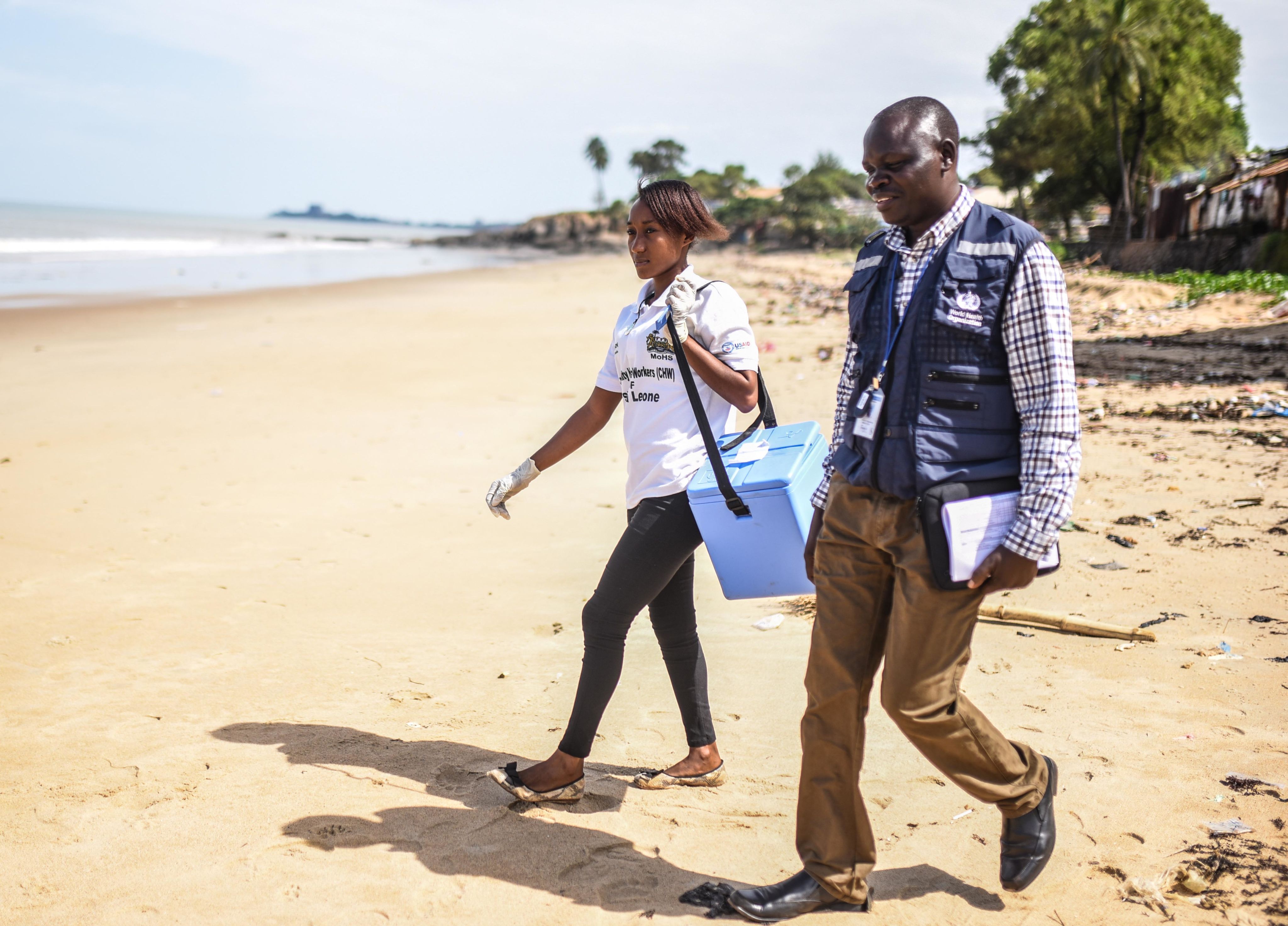 Two people walk along a beach carrying a freezer bag